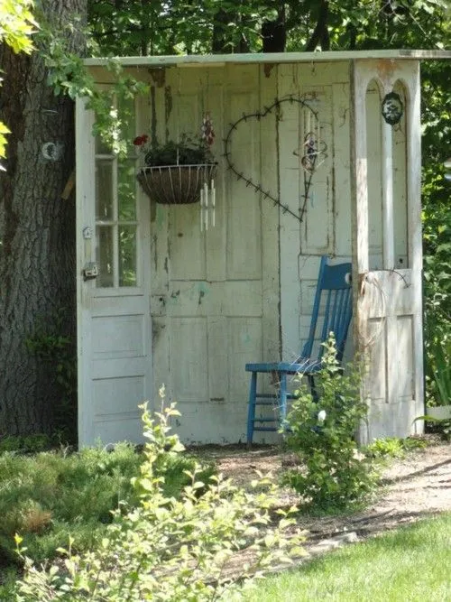 A rustic outdoor structure made from old weathered doors with a heart-shaped decoration, a hanging basket of flowers, and climbing plants